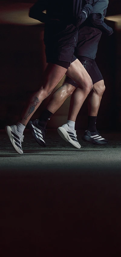 This is an action shot of two people running on what appears to be an indoor track or training area at night with artificial lighting. They are wearing dark athletic outfits and are caught mid-stride with one leg slightly ahead of the other. 