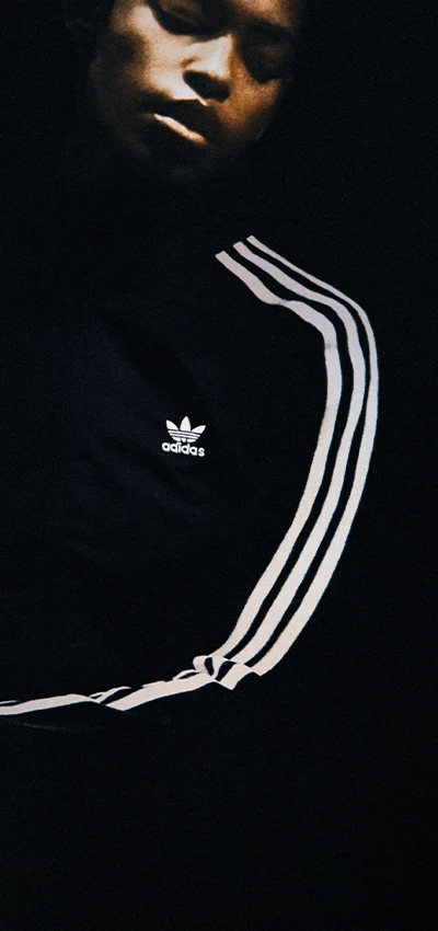 The image shows a person standing against a dark background. The individual appears to be wearing an adidas tracksuit with bold stripes and is looking downward.
