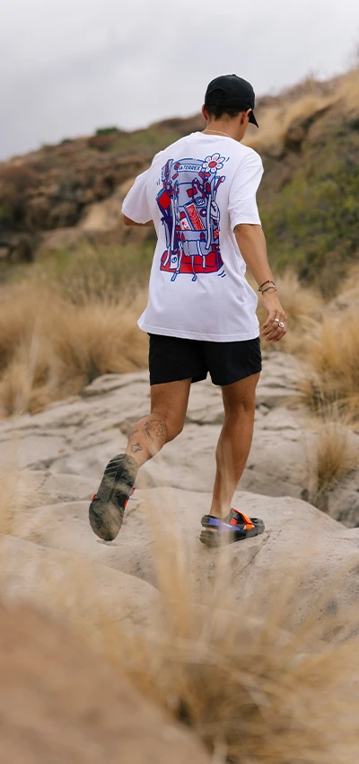 The image portrays a person wearing a white t-shirt with a graphic on the back, shorts, and sports shoes. They are positioned on some rough terrain with scrub vegetation, where they appear to be running or engaging in outdoor activity. 