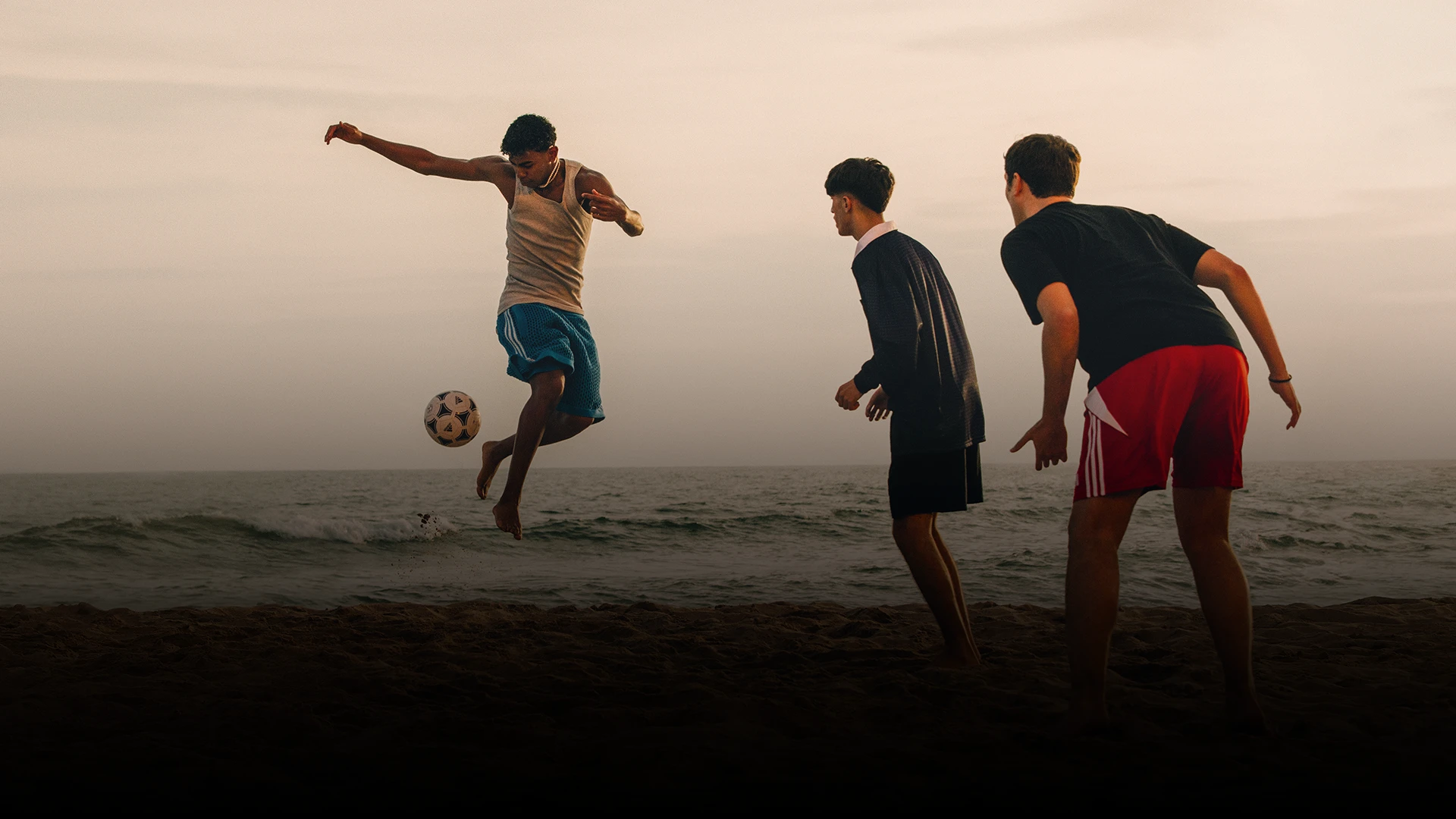 This photo shows people playing soccer on a beach at either sunrise or sunset due to the warm glow and color saturation. The image captures a moment of motion and activity, possibly from a social gathering or event involving sports.
