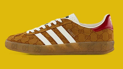 The image shows a sneaker featuring a brown, textured upper with red accents, which is characteristic of the Adidas Superstar design. The sneaker is displayed against a yellow background and has white laces contrasting with the colorful uppers.
