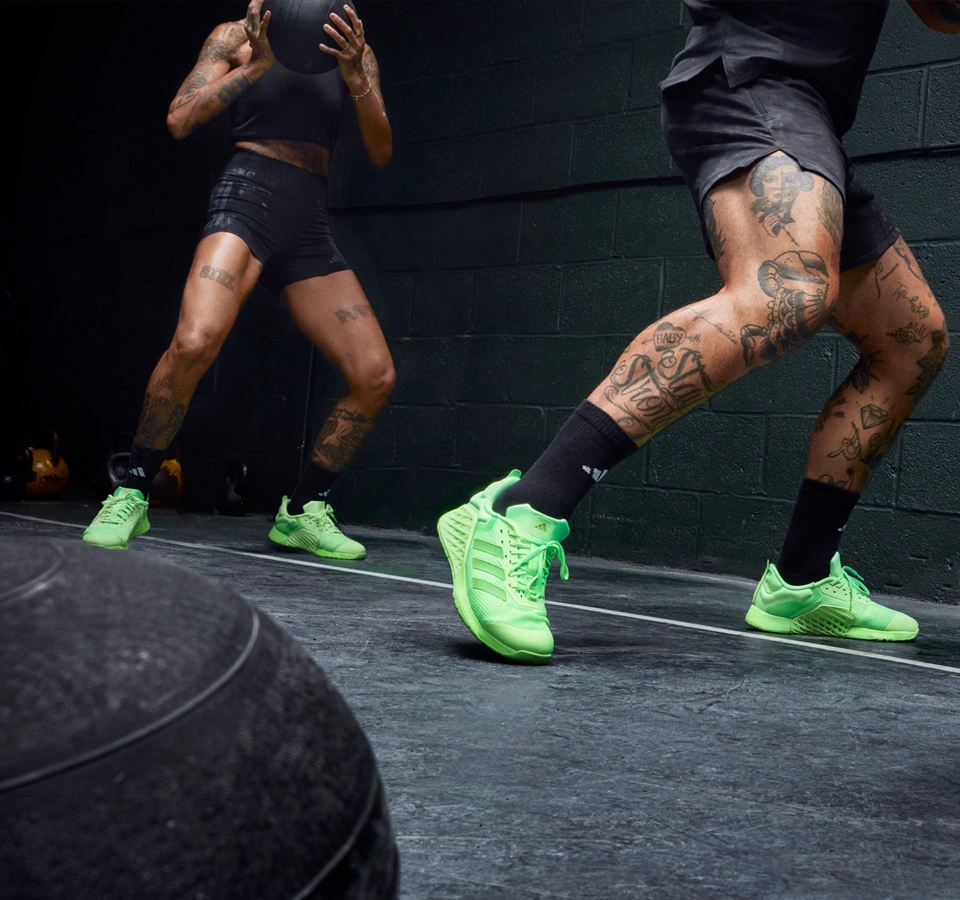Zwei Personen trainieren in einem Fitnessraum mit Medizinball und tragen neon-grüne adidas Trainingsschuhe. (Foto)