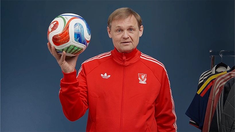 adidas CEO Bjørn Gulden im roten adidas Trainingsanzug hält einen Fußball in der Hand, neben ihm hängen Trikots an einer Kleiderstange. (Foto)