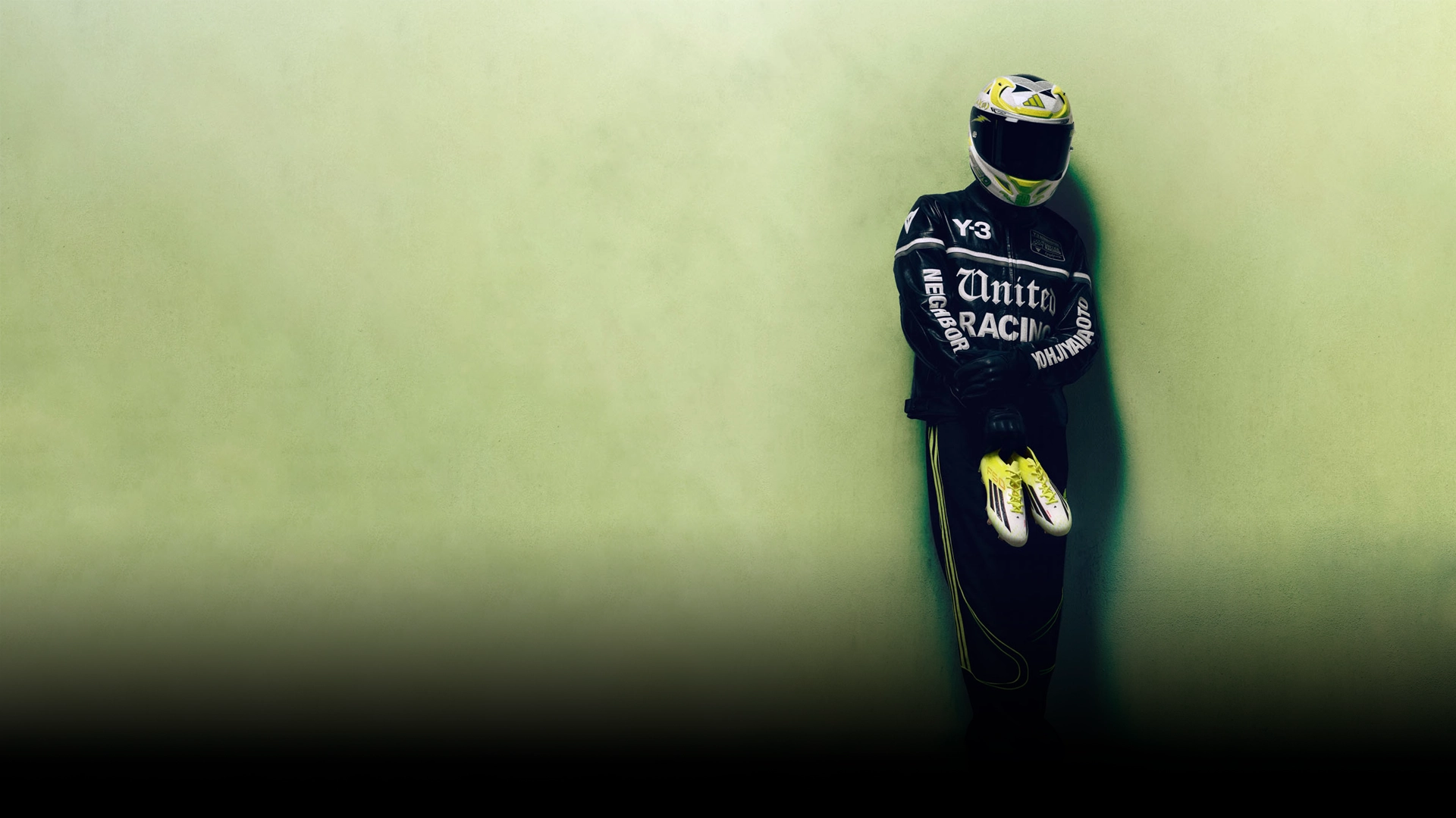 Person in a racing suit and helmet holding adidas football boots against a green background. (Photo)