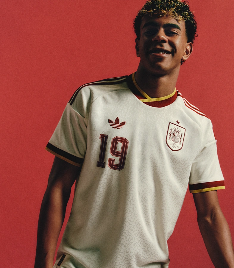 Lamine Yamal wearing a sleeveless adidas shirt, denim shorts and green adidas sneakers plays with a football against a yellow and grey wall. (Photo)