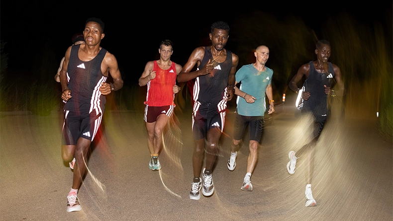 Group of five men running together at night on a road, lit by headlights. (Photo)
