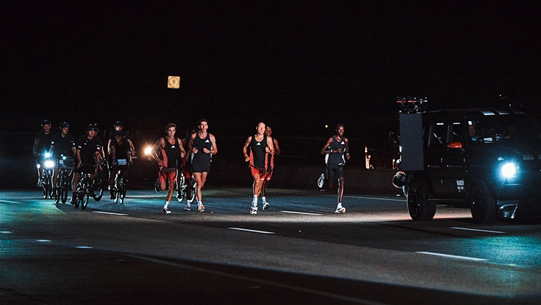 Several runners (recognizable as men) running at night on a closed road, accompanied by cyclists and a vehicle. (Photo)