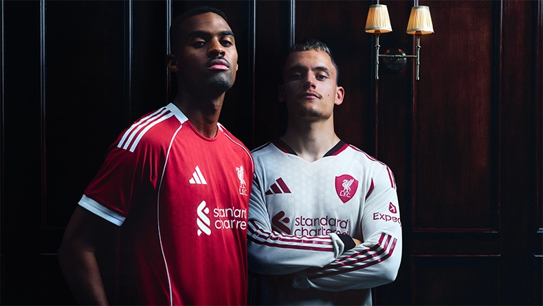 Two men wearing red and white Liverpool jerseys posing in front of dark wood paneling with wall lamps. (Photo)